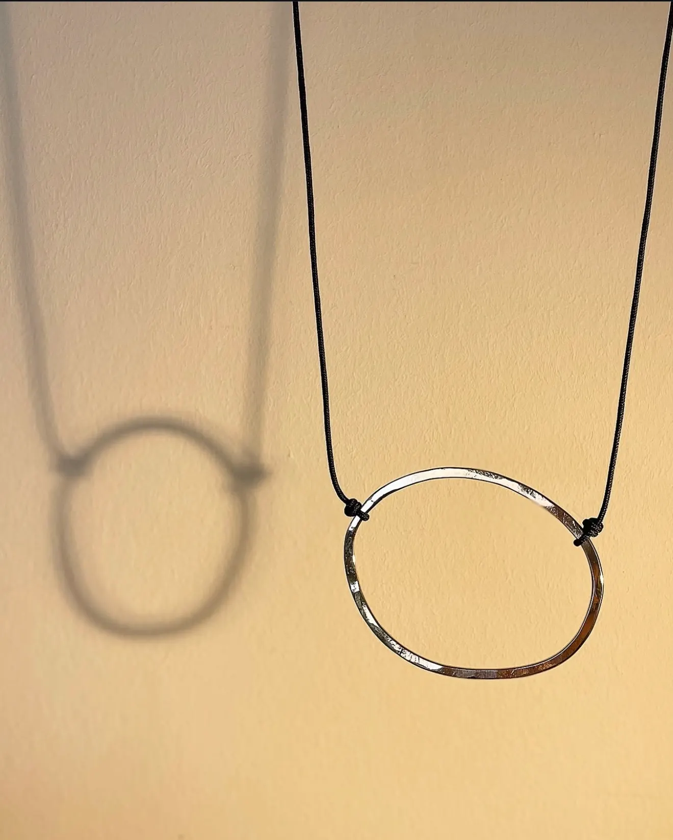 Colgante de aro martelé en plata con cordón negro, colgado sobre una pared beige con su sombra